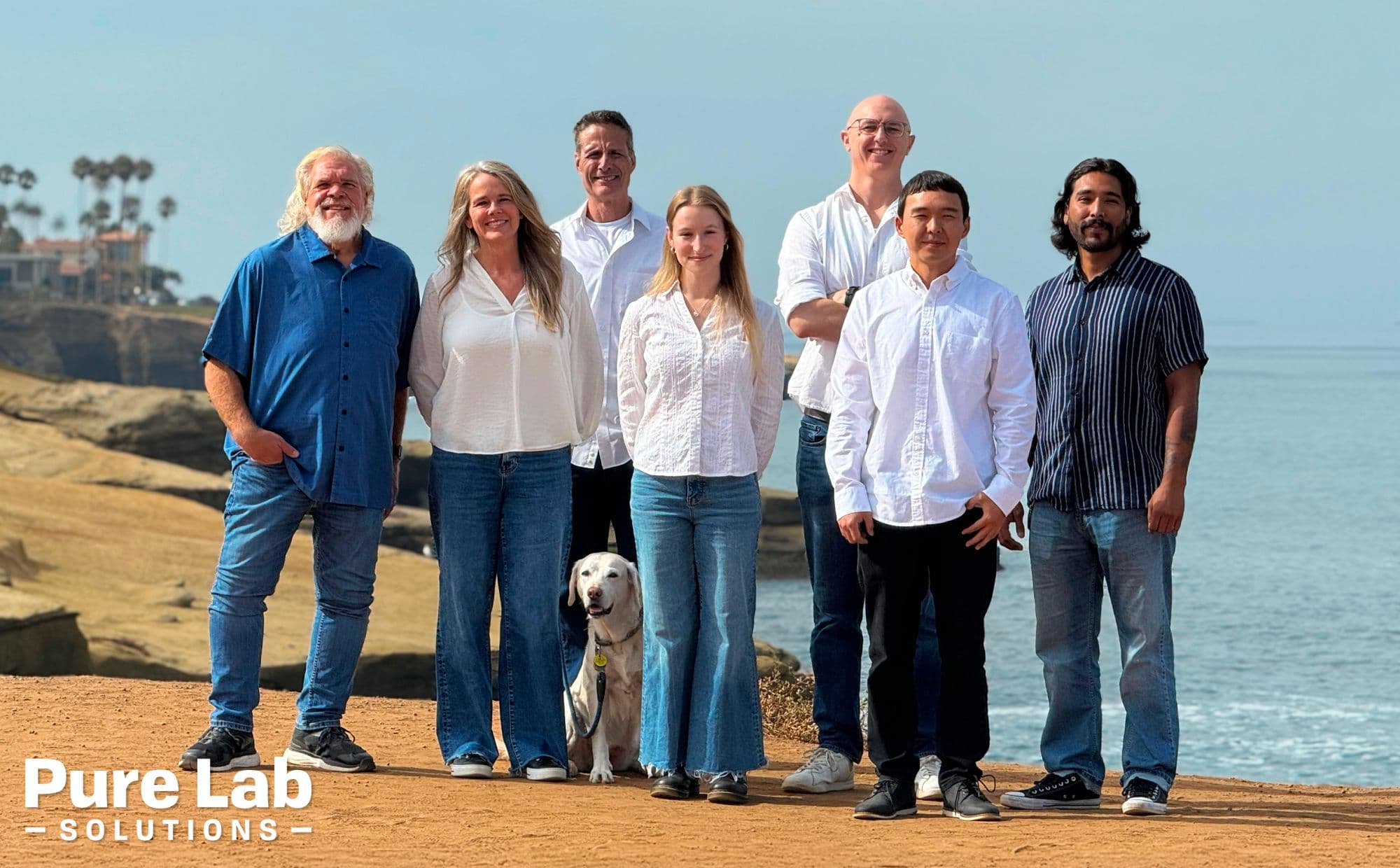 The Pure Lab Solutions team stands on a cliff, with a trailing coastline and the ocean behind them.